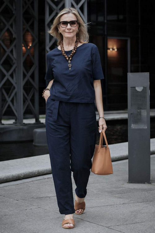 Woman wearing navy cotton linen set with short sleeves and straight-leg pants walking outdoors carrying tan handbag