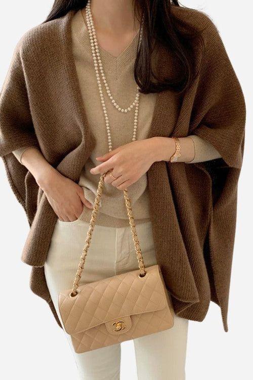 Woman wearing relaxed flowy open front brown knitwear cardigan with beige top and white pants, holding beige quilted handbag