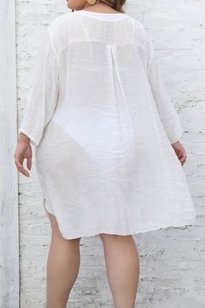 woman wearing white airy v-neck tunic with 3/4 sleeves and asymmetrical hem standing against white brick wall