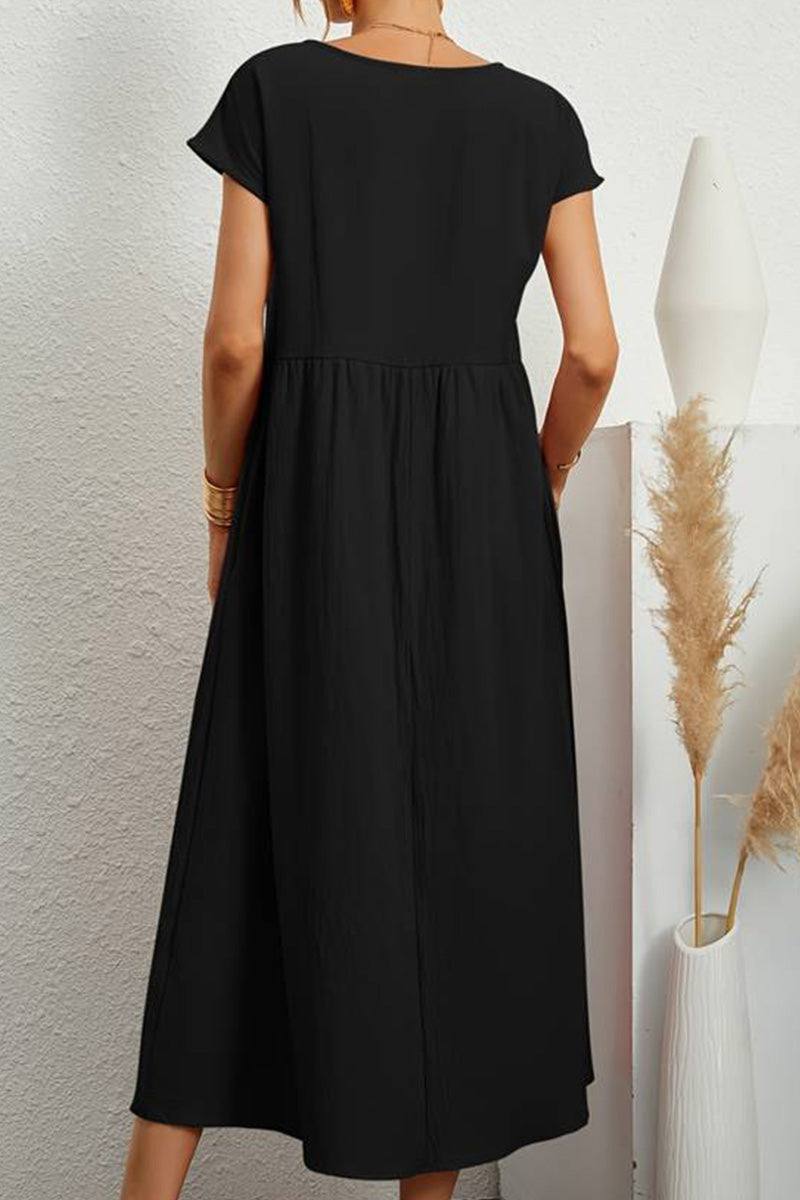 Back view of a black capped-sleeve voluminous pleated midi dress on a model against a white textured wall with decorative dried plants.