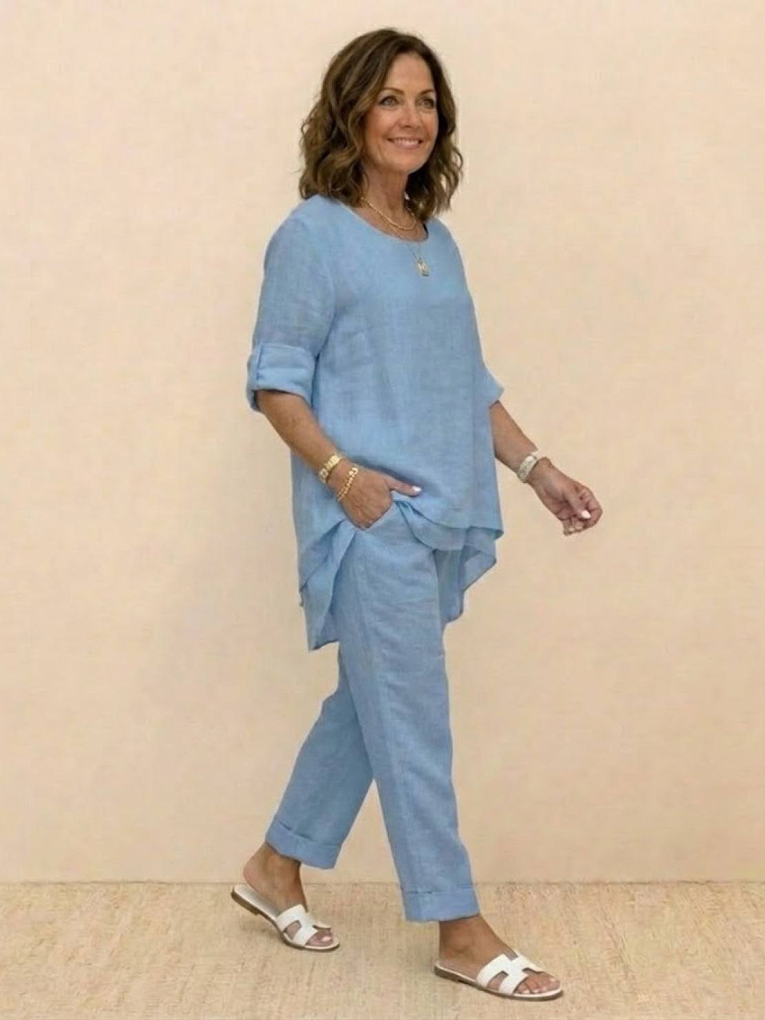 Woman wearing a light blue asymmetrical two-piece trouser set with rolled sleeves and tapered pants, styled casually with white sandals.