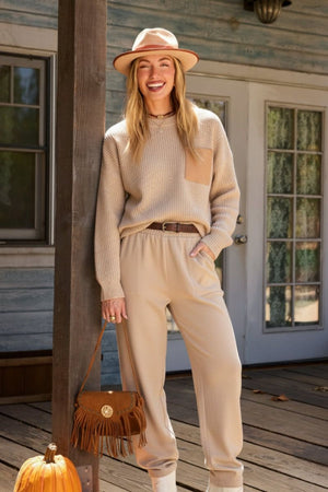 Woman wearing European minimalism relaxed sweater top and trouser set in beige standing on wooden porch with hat and handbag.