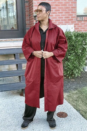 Woman wearing oversized waterproof maroon windbreaker jacket with snap-button closure and attached hood standing outdoors