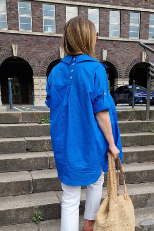 Woman wearing a bright blue oversized linen shirt with back button detailing and rolled sleeves, paired with white pants and carrying a woven bag, standing on outdoor stone steps.