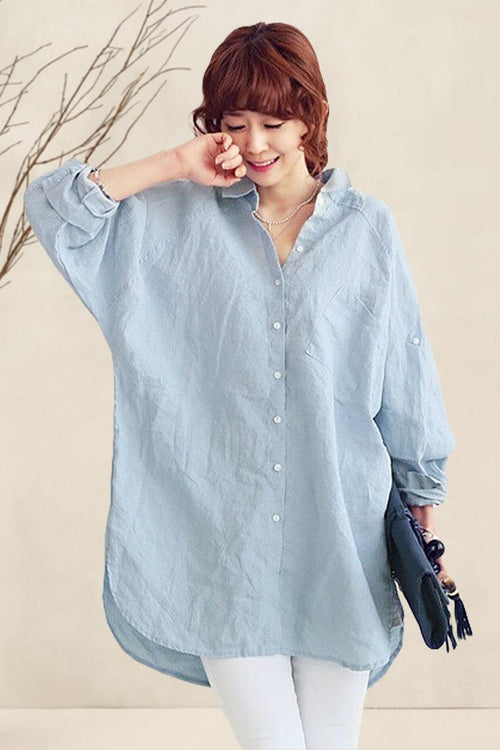 Woman wearing light blue oversized linen shirt with relaxed fit and button closure, styled casually with white pants and holding a small bag.