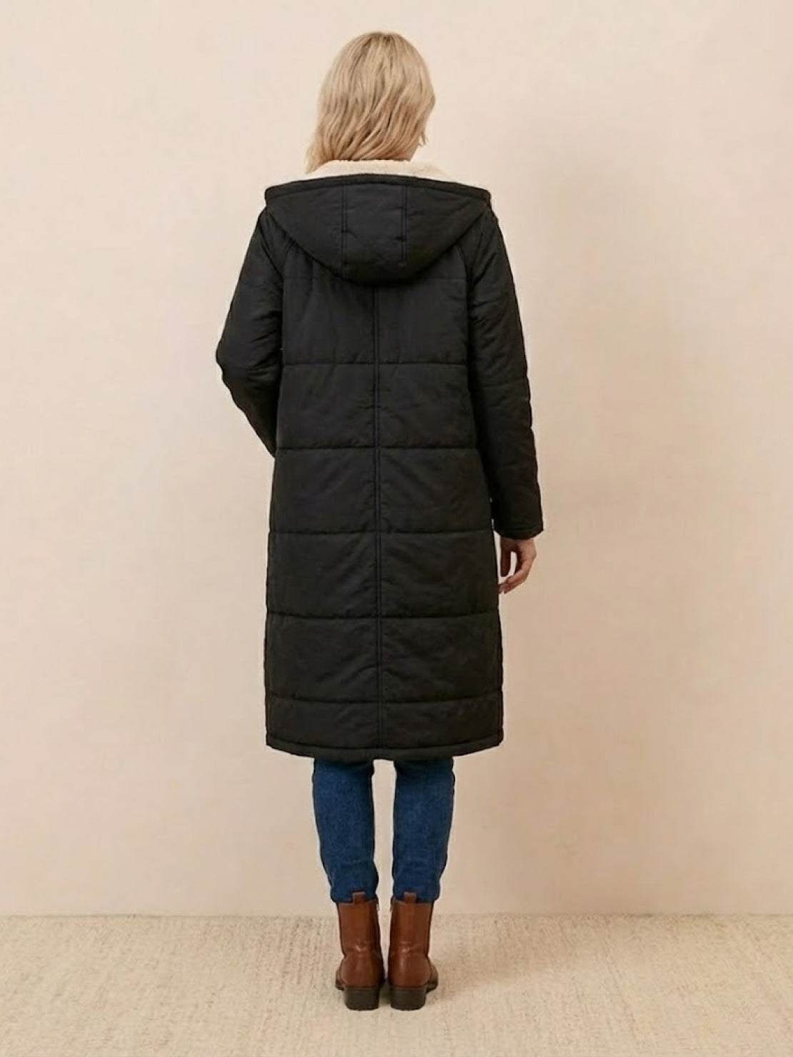 Back view of a black Sherpa-lined hooded teddy coat with an extra-long puffer design worn by a woman with blonde hair, paired with blue jeans and brown boots.
