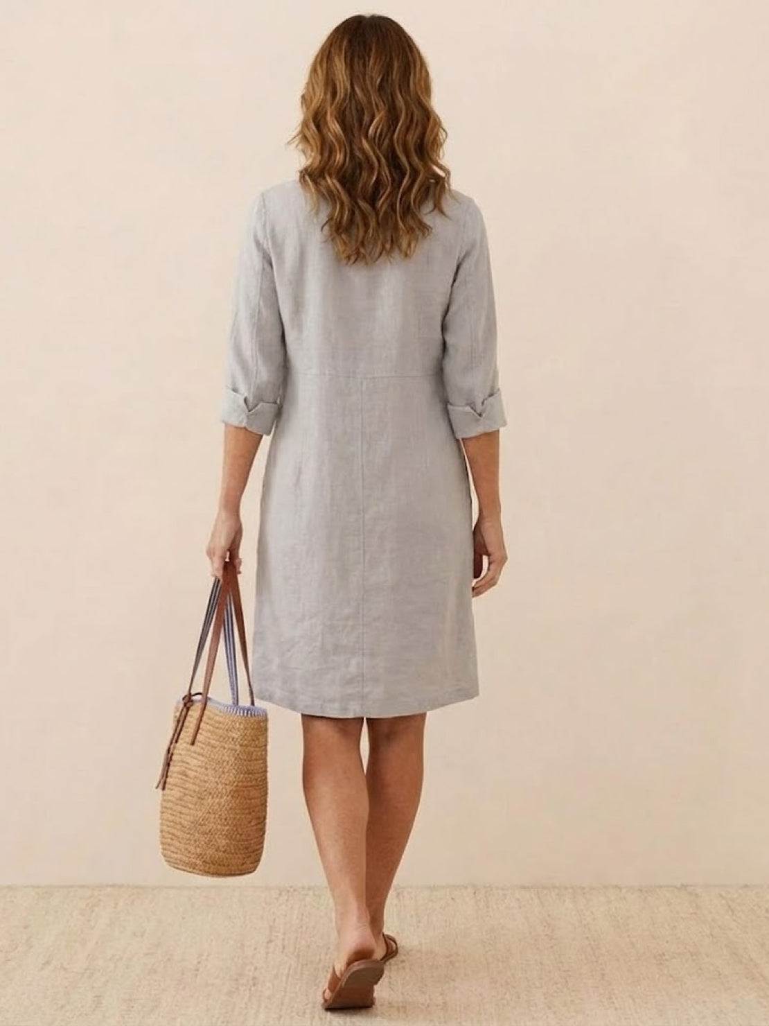 Woman wearing knee-length gray cotton dress with rolled ¾ sleeves, carrying a woven straw bag, shown from the back.