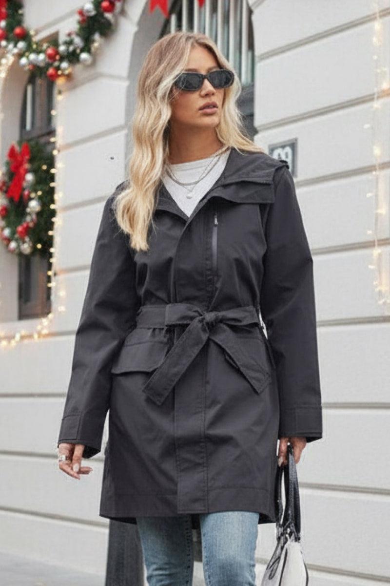 Woman wearing black water-resistant windbreaker trench jacket with belt and sunglasses outdoors