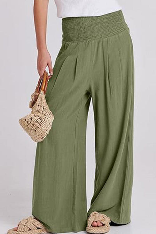 Woman wearing olive green palazzo pants with a smocked waistband holding a woven bag, paired with sandals.