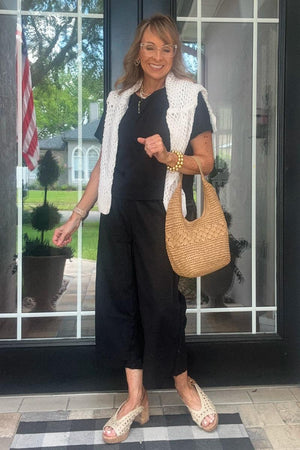 Woman wearing black loose fit co-ord set with wide-leg pants and short-sleeve top, holding woven handbag, white knit vest draped over shoulders.