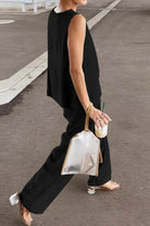 Woman wearing loose solid color black sleeveless two-piece set walking outdoors with transparent handbag and drink.