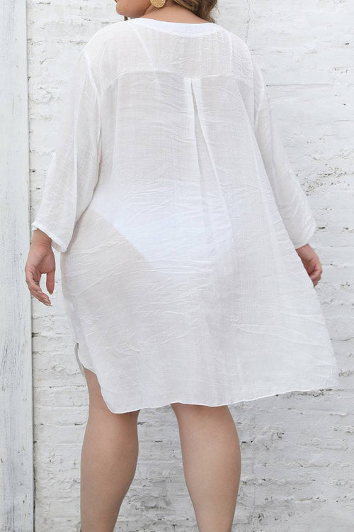 woman wearing white airy v-neck tunic with 3/4 sleeves and asymmetrical hem standing against white brick wall