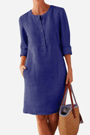 Blue cotton dress with 3/4 rolled sleeves, front buttons, and pockets, styled with a woven handbag.
