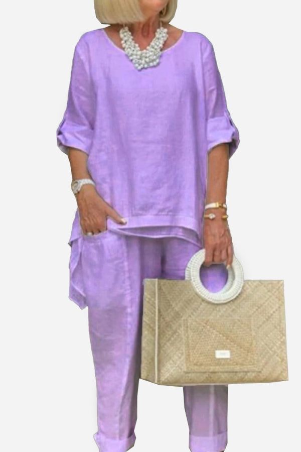 Woman wearing lavender asymmetrical two-piece trouser set with rolled-up sleeves and beige handbag