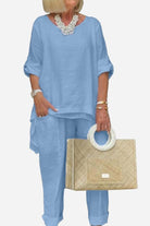 Woman wearing light blue asymmetrical two-piece trouser set with rolled sleeves and carrying beige handbag