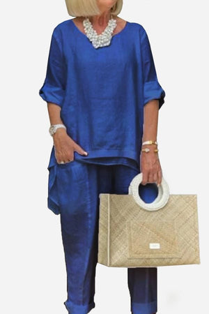 Woman wearing a royal blue asymmetrical two-piece trouser set with rolled sleeves and holding a beige woven handbag