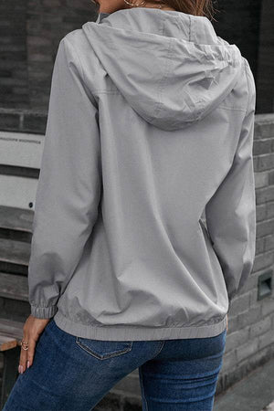 Back view of a person wearing a light grey outdoor hooded raincoat and windbreaker jacket with elastic sleeves and hem, paired with blue jeans.