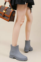 Gray non-slip rain boots with SuperGrip soles worn by person holding brown handbag.