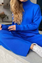 Woman wearing a bright blue knitted turtleneck two-piece set with wide-leg pants and white sneakers sitting on a ledge.