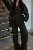 woman wearing black versatile mix-match lounge-to-travel set with wide-leg pants and belted top holding handbag indoors near stairs