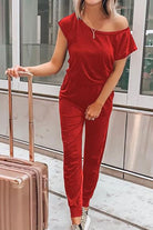 Woman wearing red Moonlit Rome Off-Shoulder Jumpsuit standing next to suitcase in modern urban setting