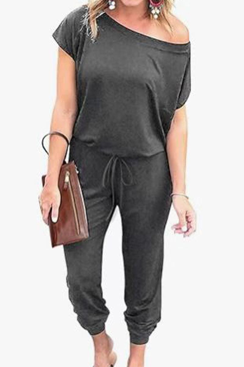 Woman wearing black Moonlit Rome Off-Shoulder jumpsuit with oblique shoulder and tapered legs, holding brown clutch.