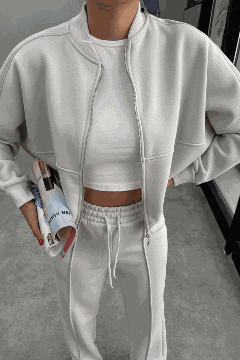 Woman wearing The Voyager Zip Set white zip-front sweatshirt and wide-leg joggers holding a magazine in a modern indoor setting