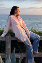 Woman wearing oversized linen shirt in soft pink, sitting by ocean at sunset
