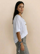 Woman wearing white pure linen age-defying shirt with button detail and rolled sleeves, paired with gray pants.