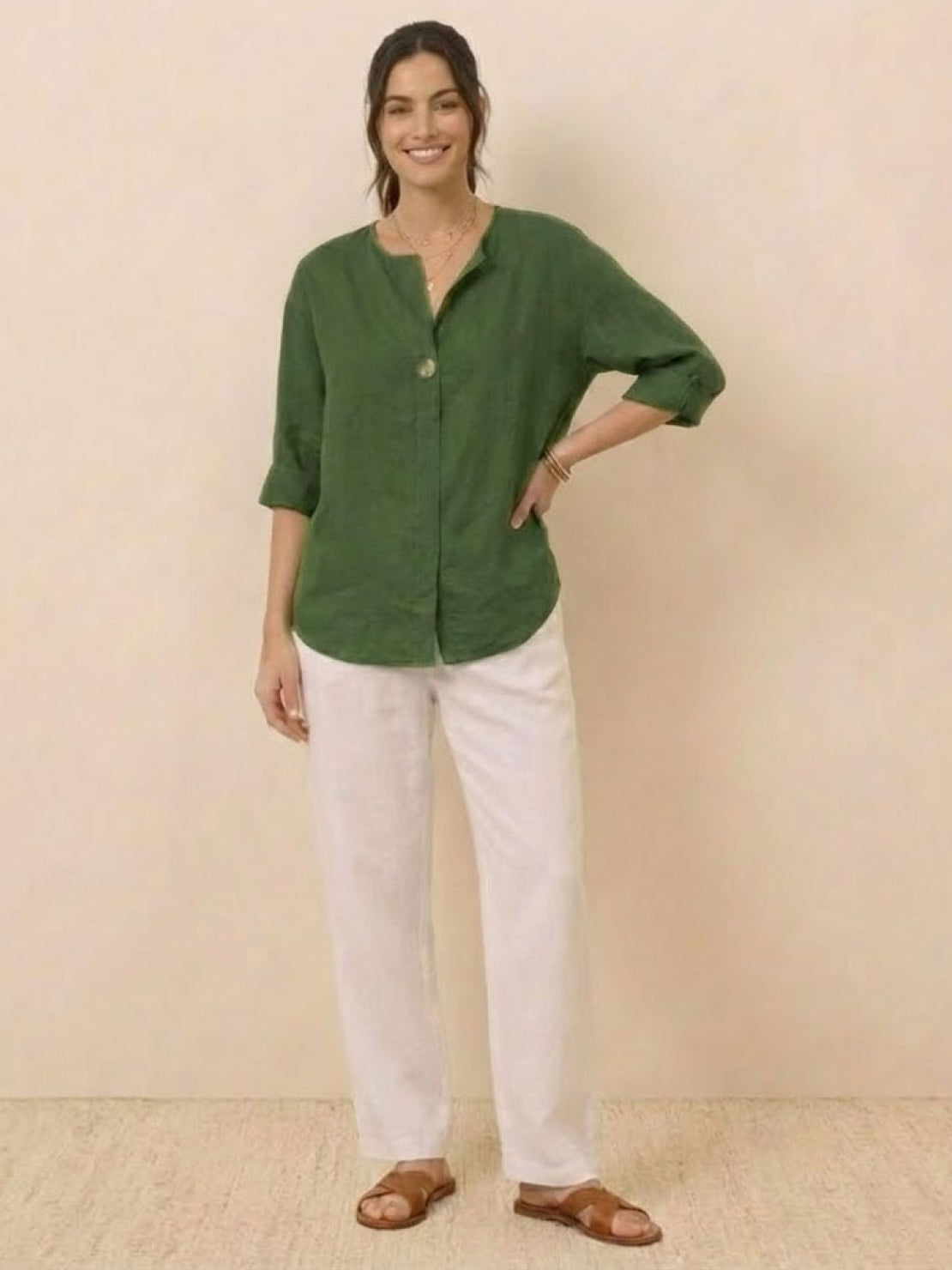 woman wearing green pure linen age-defying button detail shirt with white pants and sandals standing against beige background