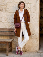 woman wearing brown Rain Couture Windbreaker with white pants and maroon sneakers outdoors against stone wall