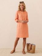 Woman wearing coral cotton dress with 3/4 sleeves and button detail, styled with sandals and a straw bag, standing against a neutral background