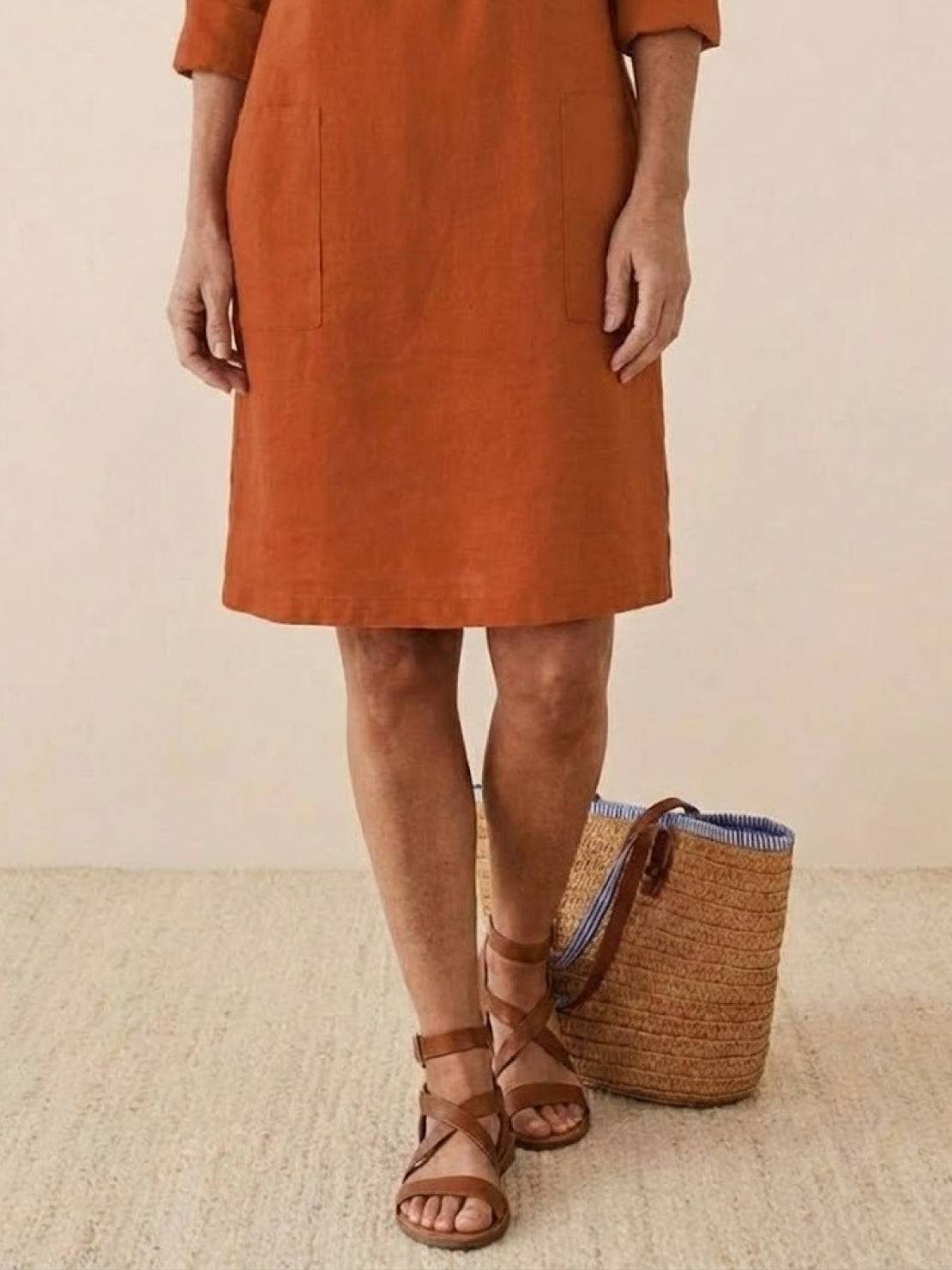 Women's cotton dress in earthy brown with knee-length hem and pockets, paired with brown strappy sandals and a woven basket bag.